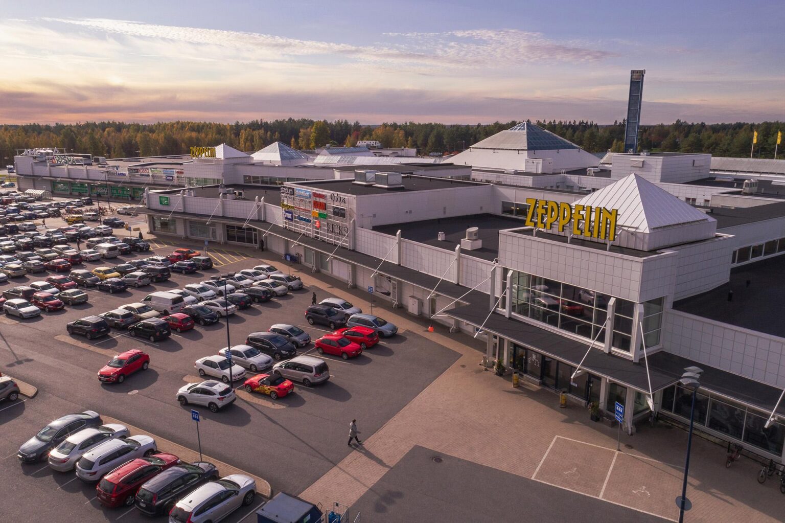 Ilmakuva Zeppelin-kauppakeskuksesta Kempeleessä osana Spondan case-esittelyä. Kuvassa näkyy rakennuksen arkkitehtuuri, laaja parkkialue ja ympäröivää metsää ilta-auringon valossa.
