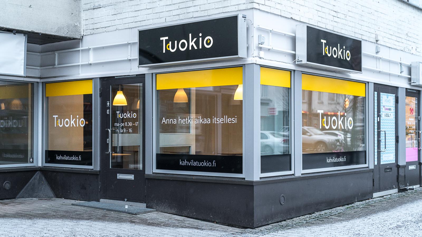 Kahvila Tuokion yritysilmeen mukainen liiketilan julkisivu, jossa näkyvät kelta-mustat ikkunateippaukset, "Anna hetki aikaa itsellesi" -slogan sekä valokyltit Luma Creativen suunnittelemalla logolla.